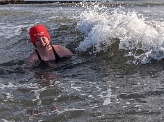Photo of a woman in the sea. She is wearing a bright red Irish Heart Foundation beanie hat and has a joyful, shocked look on her face as a wave crashes beside her