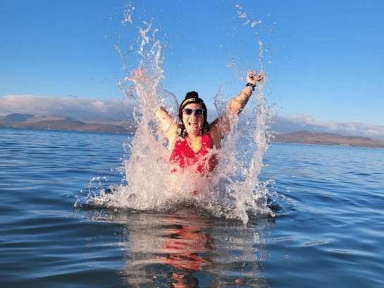 Photo of a woman seemingly emerging from the sea water as the water splashes up around her. She is wearing sunglasses and a red swimming costume. It is a beautiful day with blue skies behind her.