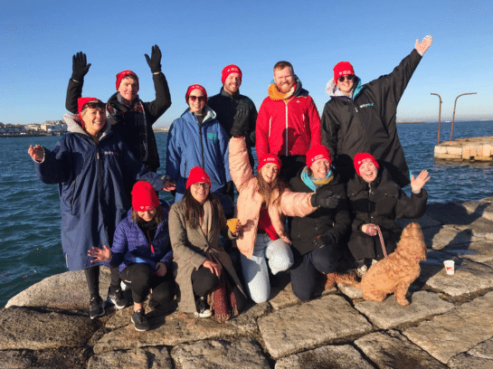 Photo of a group of people standing on the water's edge. Some are standing in a row with their arms raised in a wave whilst the others kneel in front, smiling at the camera. Blue skies behind them.