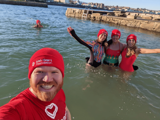 Photo taken selfie style by a man, Jordan, wearing a red Irish Heart Foundation t-shirt and beanie hat. He is in the water and behind him are three people also in the water. They have their arms around each other and are waving at the camera. They too wear the red beanie hats.