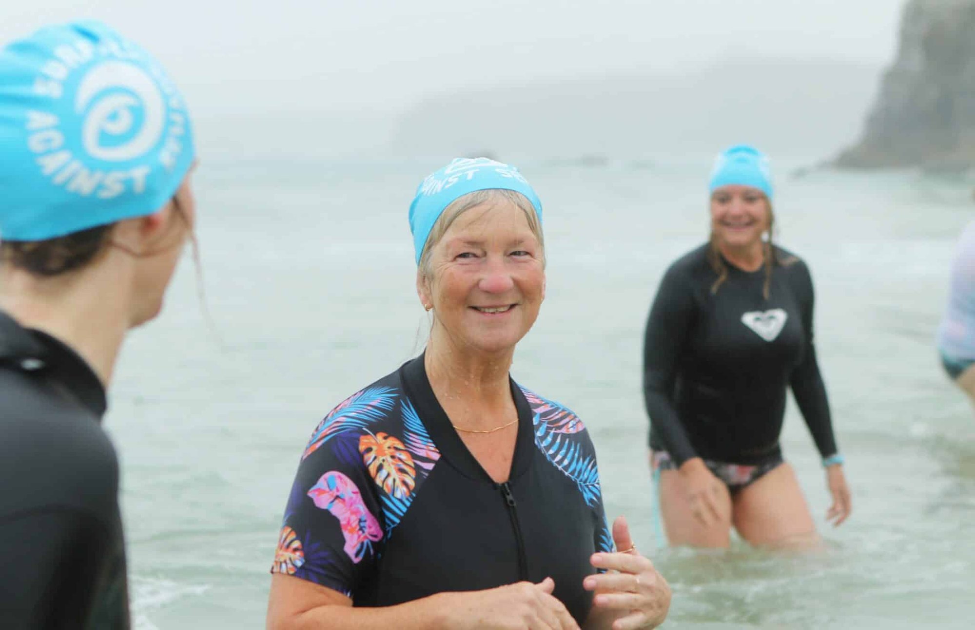 Photo of women standing in the sea. One woman is facing the camera, smiling. They are all wearing light blue swim caps which have Surfers Against Sewage's logo on the side. It is misty and the water is cloudy.