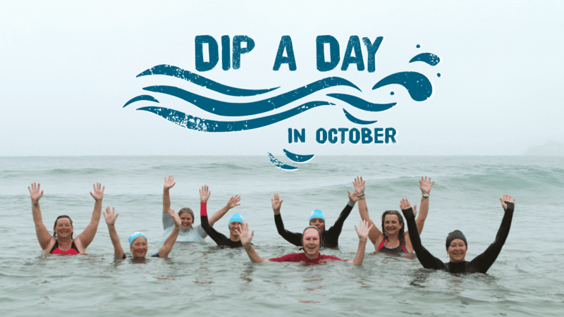 Photo of eight women in the sea with the water up to their shoulders. They all have their arms raised in the air and are smiling at the camera. They all wear light blue swim caps with Surfers Against Sewage's logo on the side. Above them is a text overlay of Dip a Day in October with a wave design running underneath.