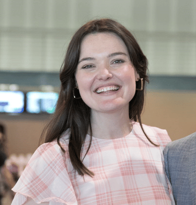 Photo of Michaela Britt. She is a young woman with shoulder-length brown hair and is wearing a pink and white checked top. She is smiling towards the camera.