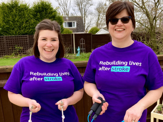 Photo of two women outside; they both wear purple Stroke Association t-shirts which has the text 'Rebuilding lives after stroke' on the front. They are both holding skipping ropes and smiling at the camera.