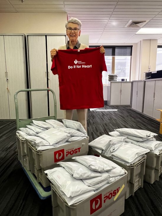 Photo of a Heart Foundation volunteer, an older woman with short hair and glasses. She is holding up a Heart Foundation red t-shirt and is standing infant of boxes of packaged t-shirts that are ready to send.